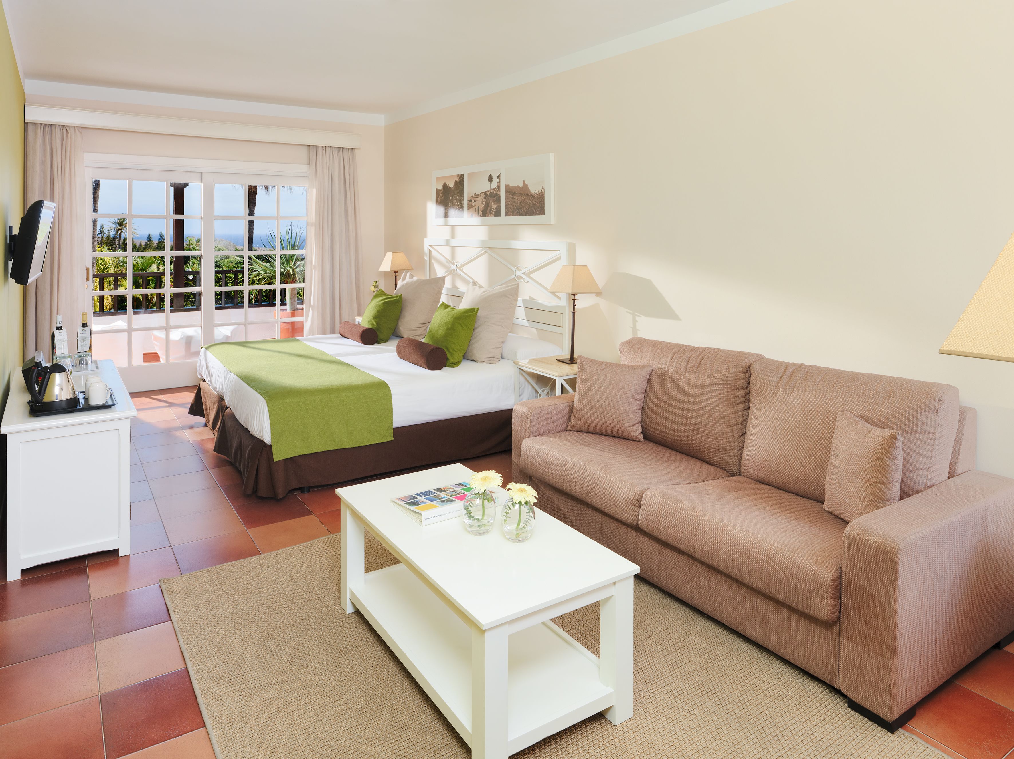 A hotel room with a bed, a sofa, a coffee table, a desk with a lamp, and a view of the outdoors.