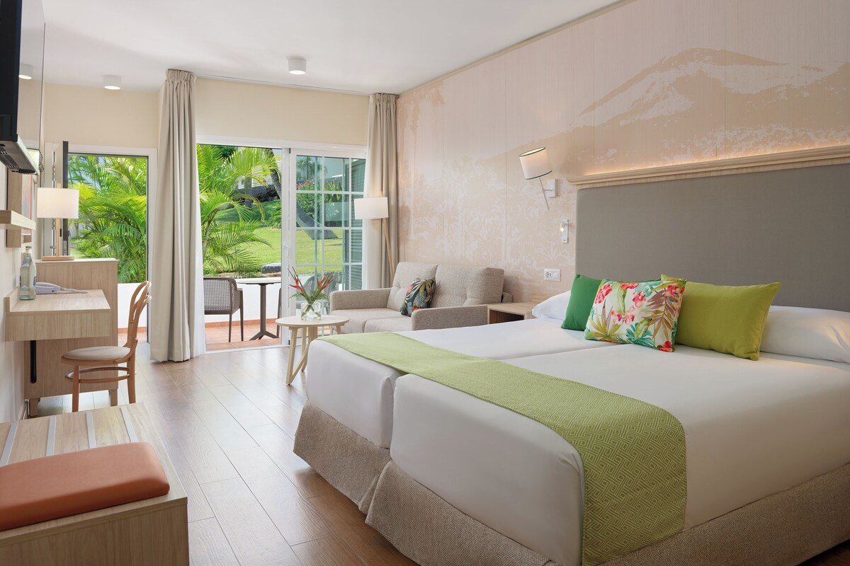 A hotel room with a bed, two bedside tables, a desk, and a chair. There is a view of greenery outside the window.
