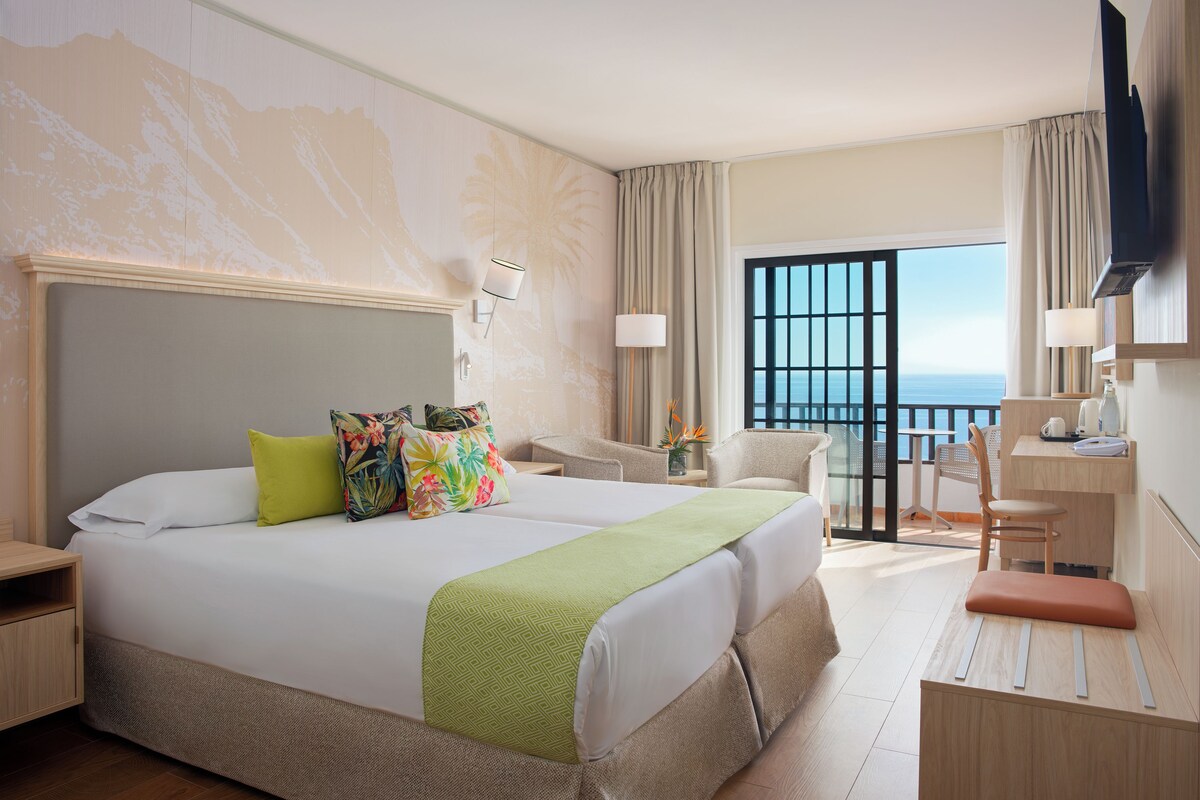 A hotel room with a large bed, a desk, a chair, a TV, and a balcony with a view of the sea.