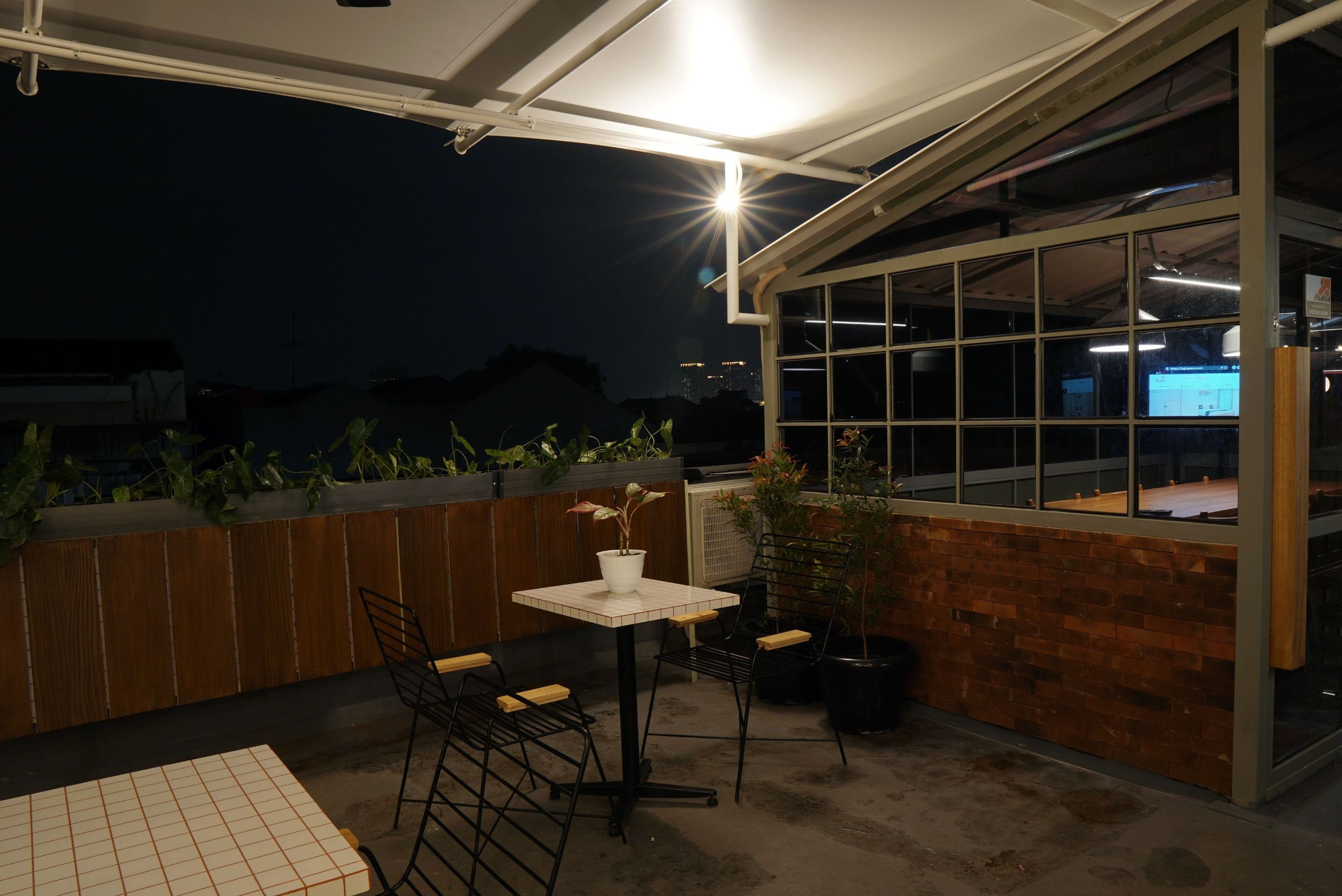 An outdoor patio area with a table and chairs, a potted plant, and a view of the cityscape at night.