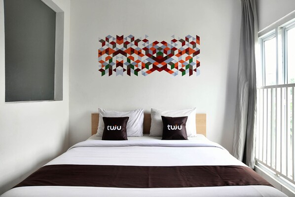 A hotel room with a bed, two pillows with the brand name 'tuju', a geometric wall art piece, and a window with curtains. - Tuju Abuserin Syariah (Jakarta)