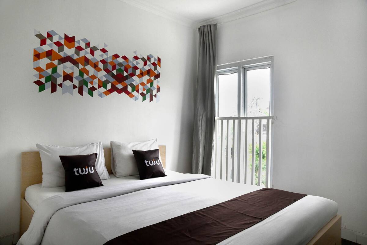 A hotel room with a bed, two pillows, a geometric wall art piece, a window with curtains, and a balcony view.
