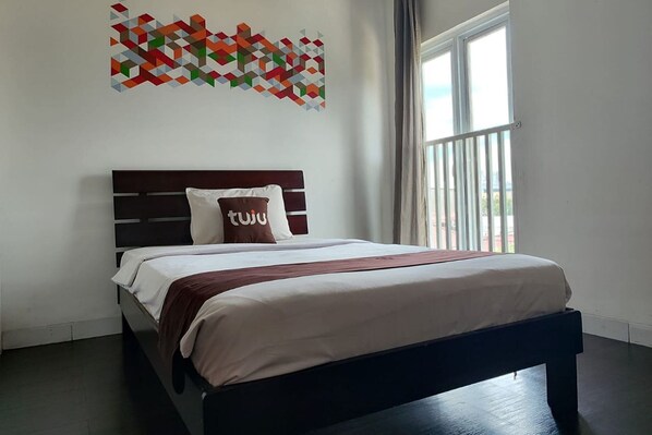 A neatly made bed with a dark headboard, a geometric wall art piece above it, and a large window with curtains to the right. - Tuju Abuserin Syariah (Jakarta)