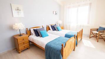 A room with two wooden beds, a wooden nightstand, a chair, and a window with sheer curtains.