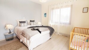 A bedroom with a bed, a bedside table, a lamp, a crib, and a window with curtains. - Villa Mojito (Pegeia)
