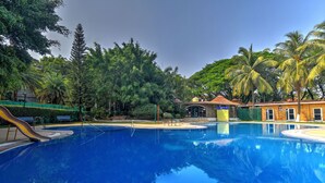 Une piscine entourée d’une végétation luxuriante et de palmiers, avec un petit pont et un bâtiment à toit de tuiles en arrière-plan.