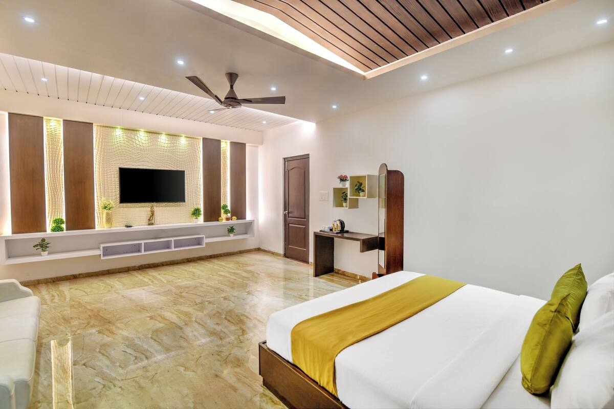 A modern hotel room with a large bed, a flat-screen TV mounted on the wall, a ceiling fan, and a wooden ceiling.