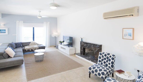 A modern living room with a grey sofa, a patterned armchair, a flat-screen TV, a fireplace, and an air conditioning unit.