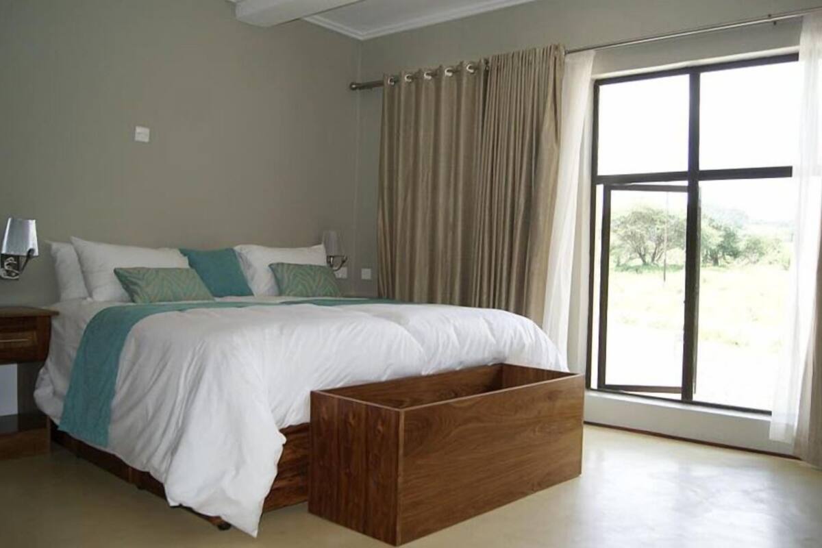 A bedroom with a large bed, a wooden bench, a nightstand, a lamp, and a window with curtains.