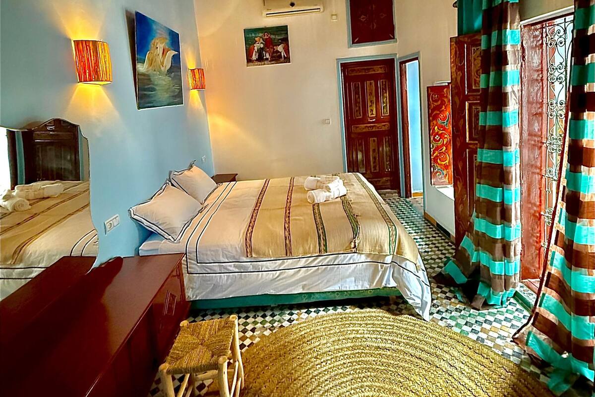 A bedroom with a bed, bedside tables, a desk, a chair, a woven rug, a hanging light fixture, and a window with curtains.