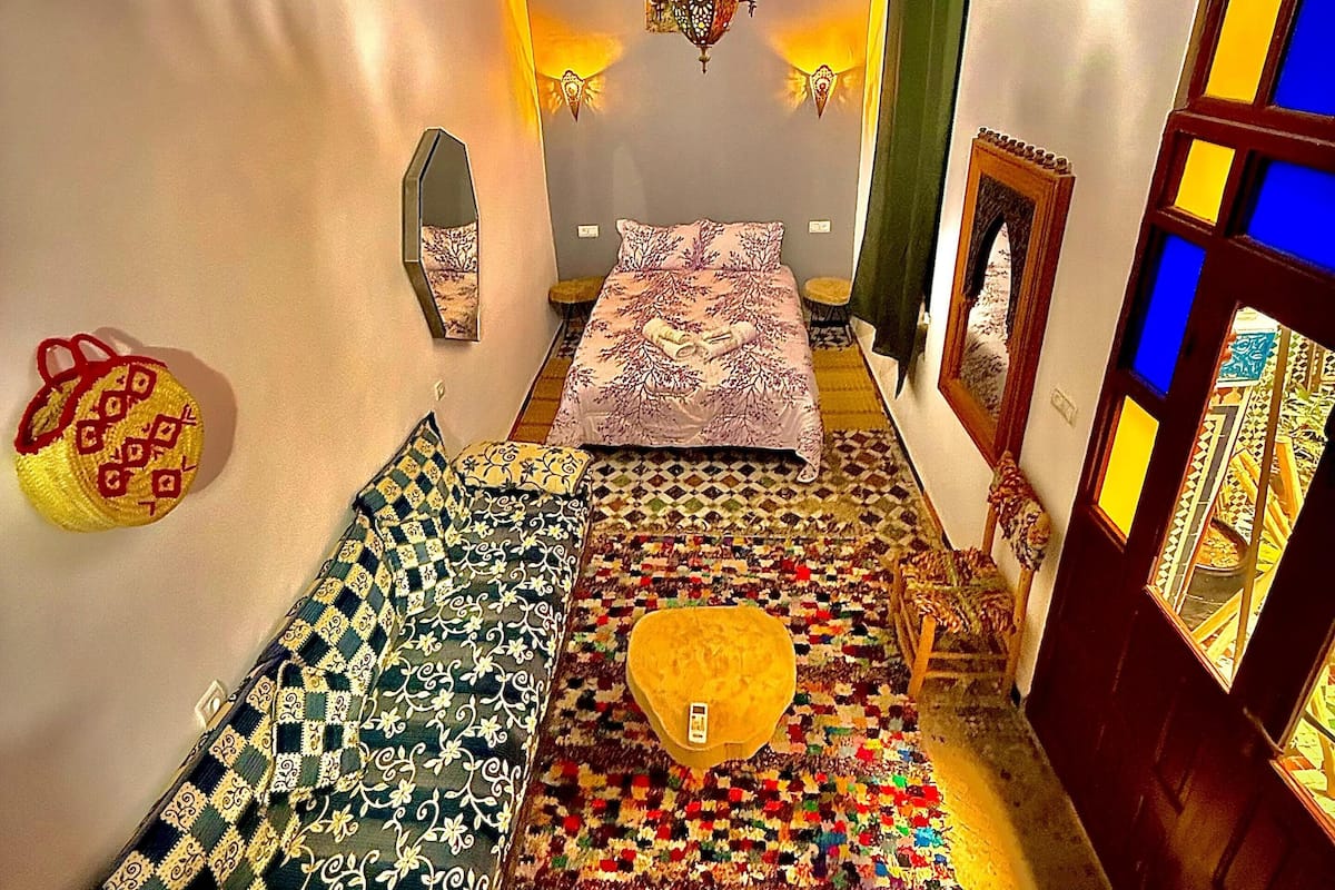 A room with a bed, two sofas, a colorful rug, a hanging light fixture, and a door with stained glass.