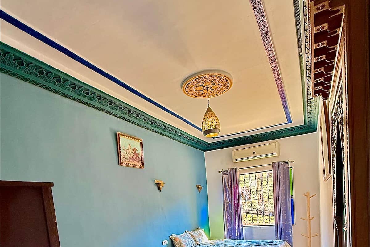 A room with two beds, a colorful rug, a decorative ceiling, and a window with curtains.
