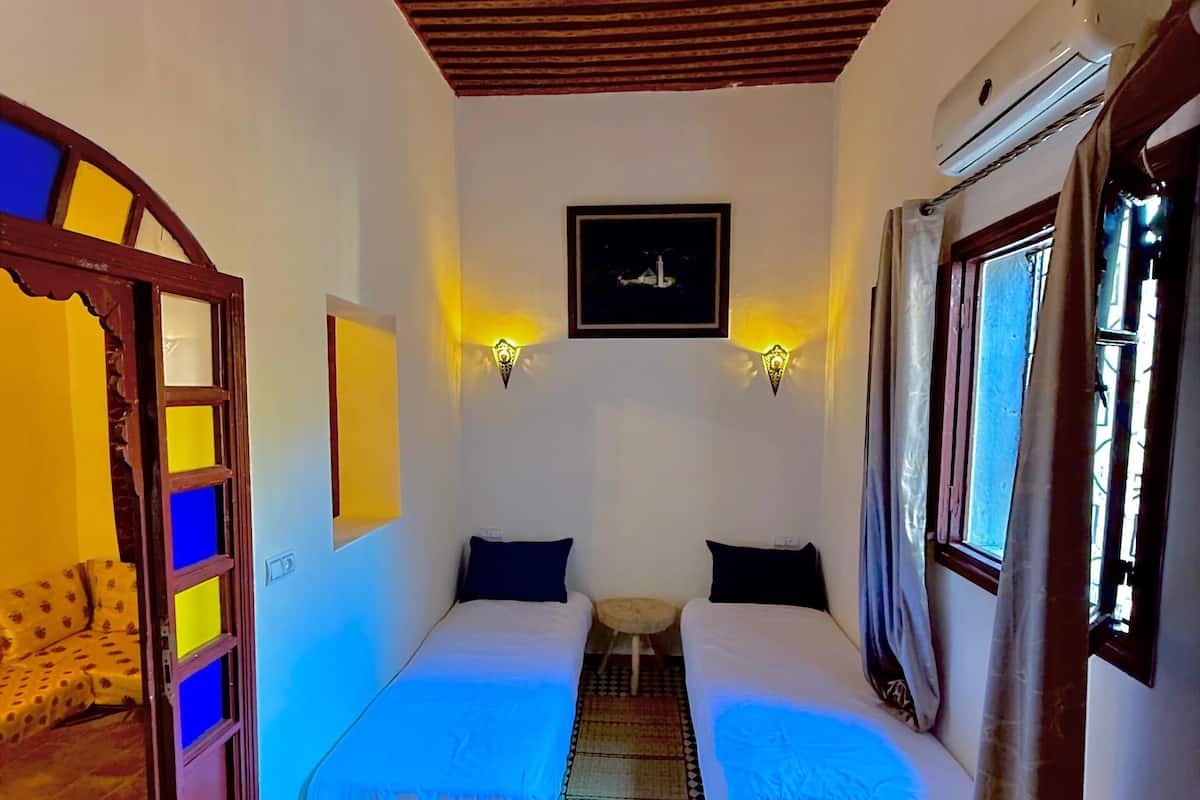 A room with two beds, a small table, a ceiling light, and a window with curtains.
