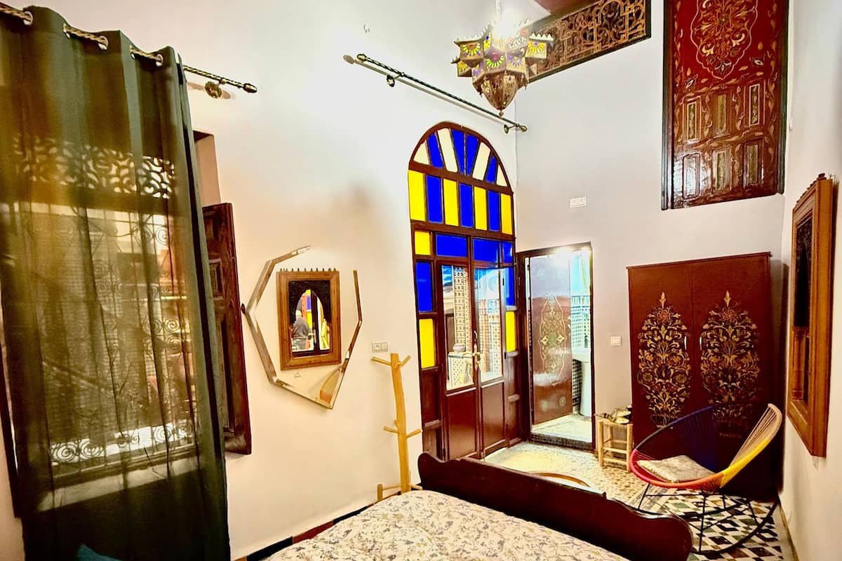 A bedroom with a bed, a chair, a window with blue and yellow panes, and a decorative wall panel.