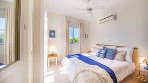 A bedroom with a bed, a ceiling fan, an air conditioning unit, a bedside table with a lamp, and a window with curtains. - Villa Porto (Pegeia)