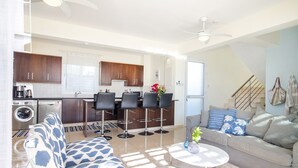 A modern apartment with a living room, kitchen, and dining area. It includes a sofa, dining chairs, a coffee table, and a kitchen with a refrigerator, stove, and sink. - Villa Porto (Pegeia)