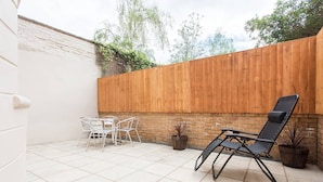 A patio with a white tiled floor, a wooden fence, a black lounge chair, and two white metal chairs. - The New Kent Road Apartments (London)