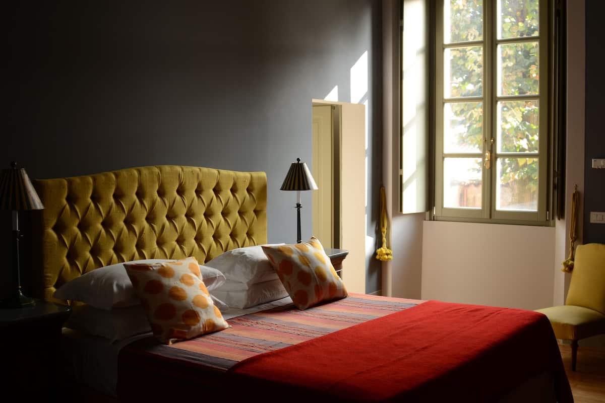 Ein Schlafzimmer mit einer gesteppten Kopfteil, einem Bett mit gemusterten Kissen, einer roten Bettdecke, einem Nachttisch mit Lampe und einem Fenster mit Blick auf Grünflächen.