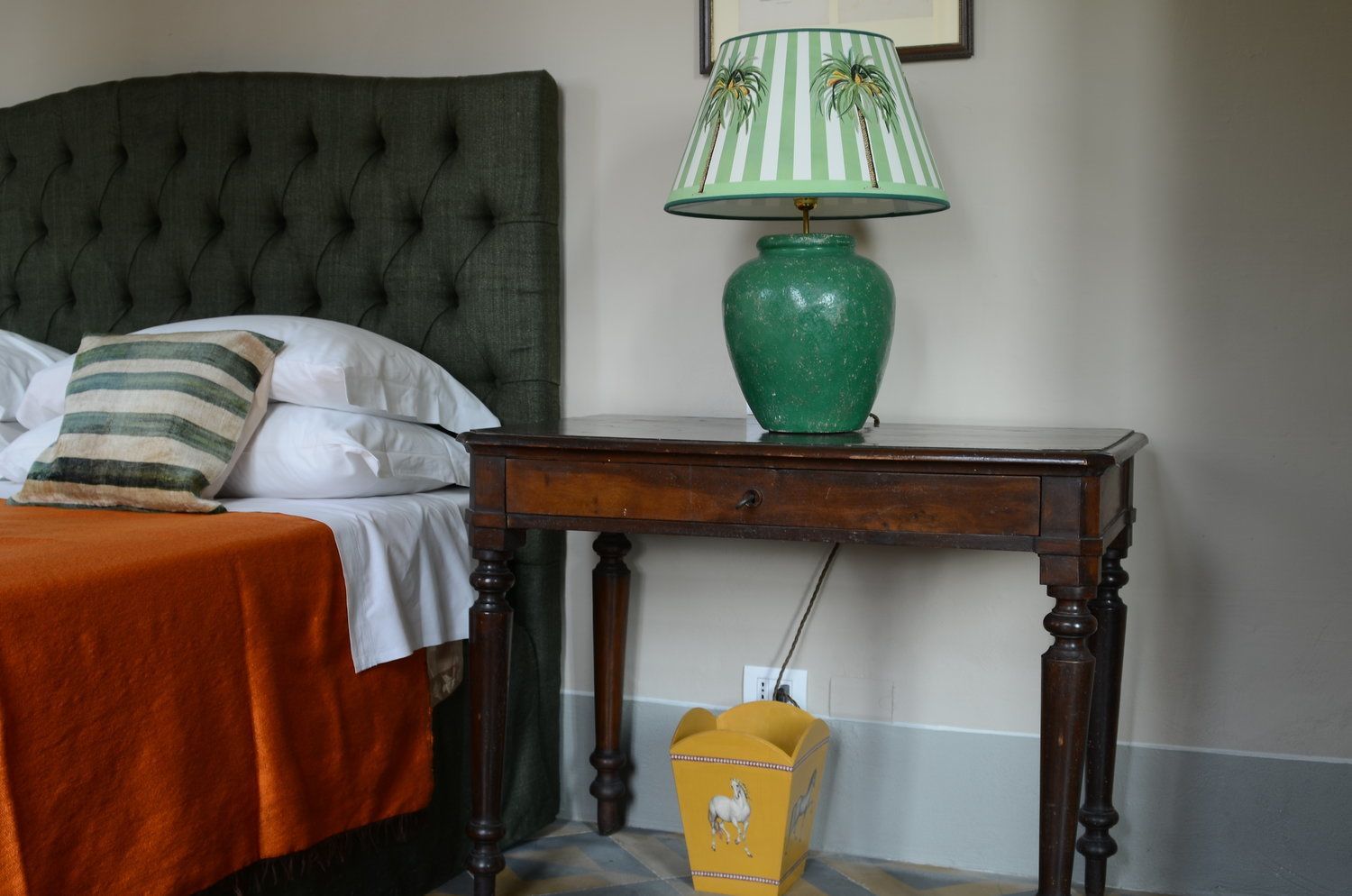 Una cama bien hecha con un cojín a rayas, una lámpara verde sobre una mesita de noche de madera y una caja amarilla con un diseño de caballo.