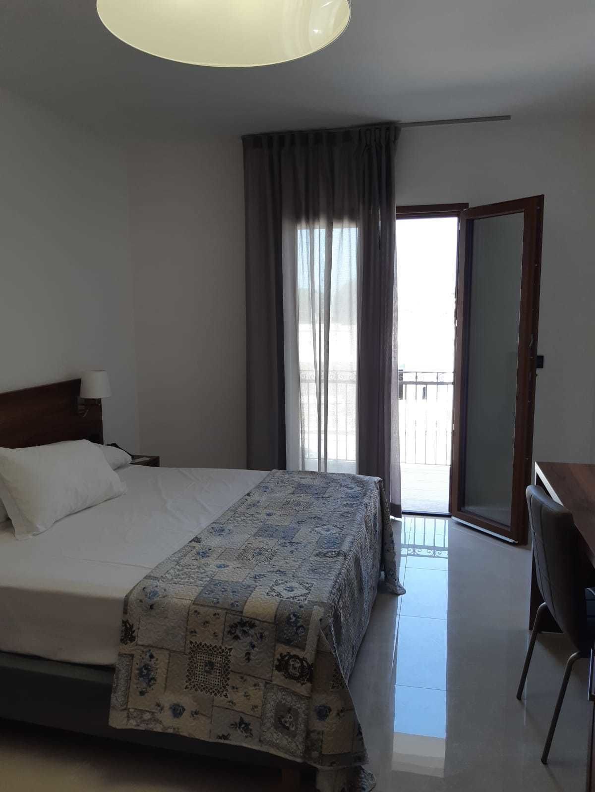 Quarto de hotel com cama, escrivaninha, cadeira, janela grande com cortinas e varanda.