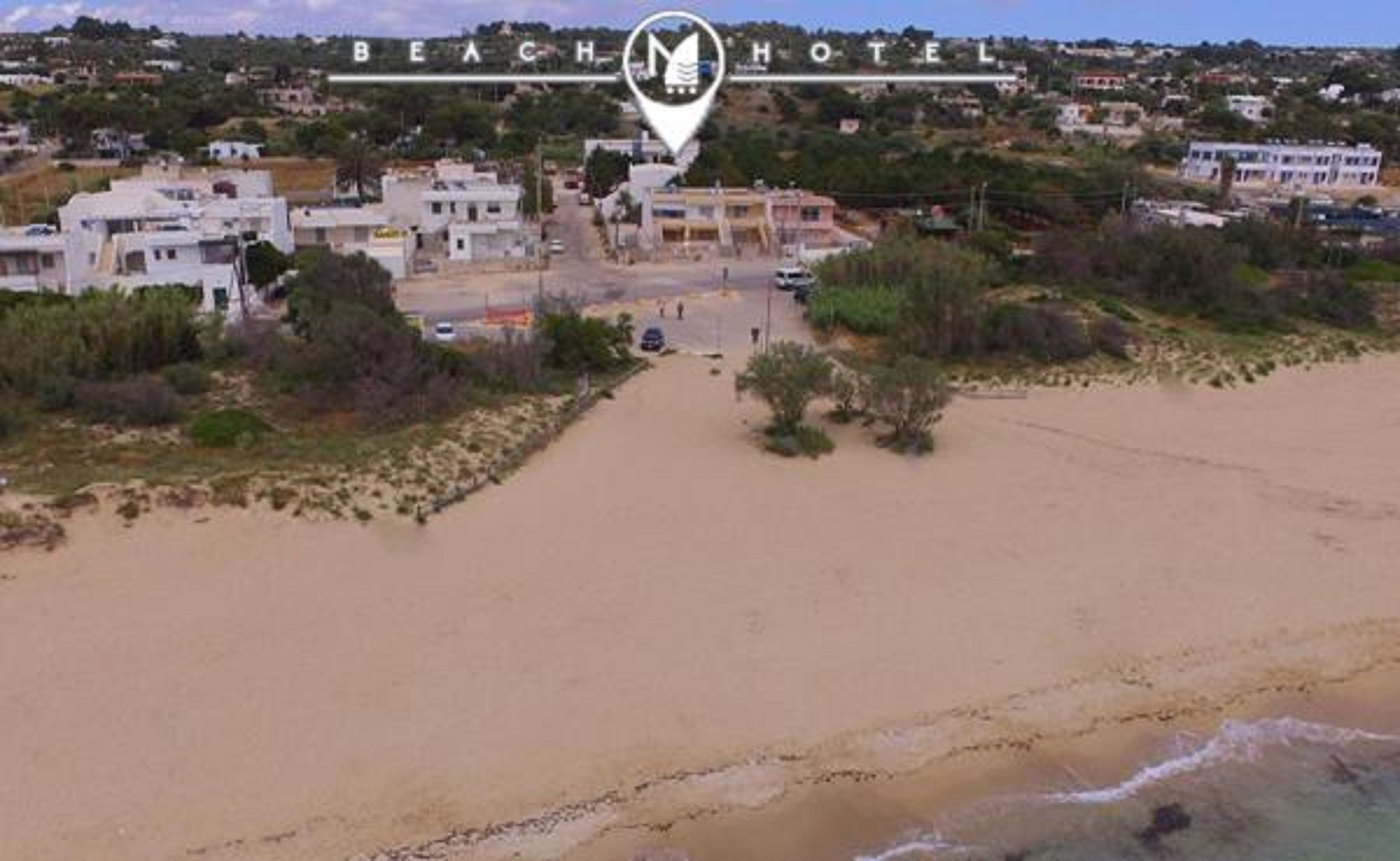 Un hôtel situé en bord de mer, avec une plage de sable, un parking et des bâtiments en arrière-plan.