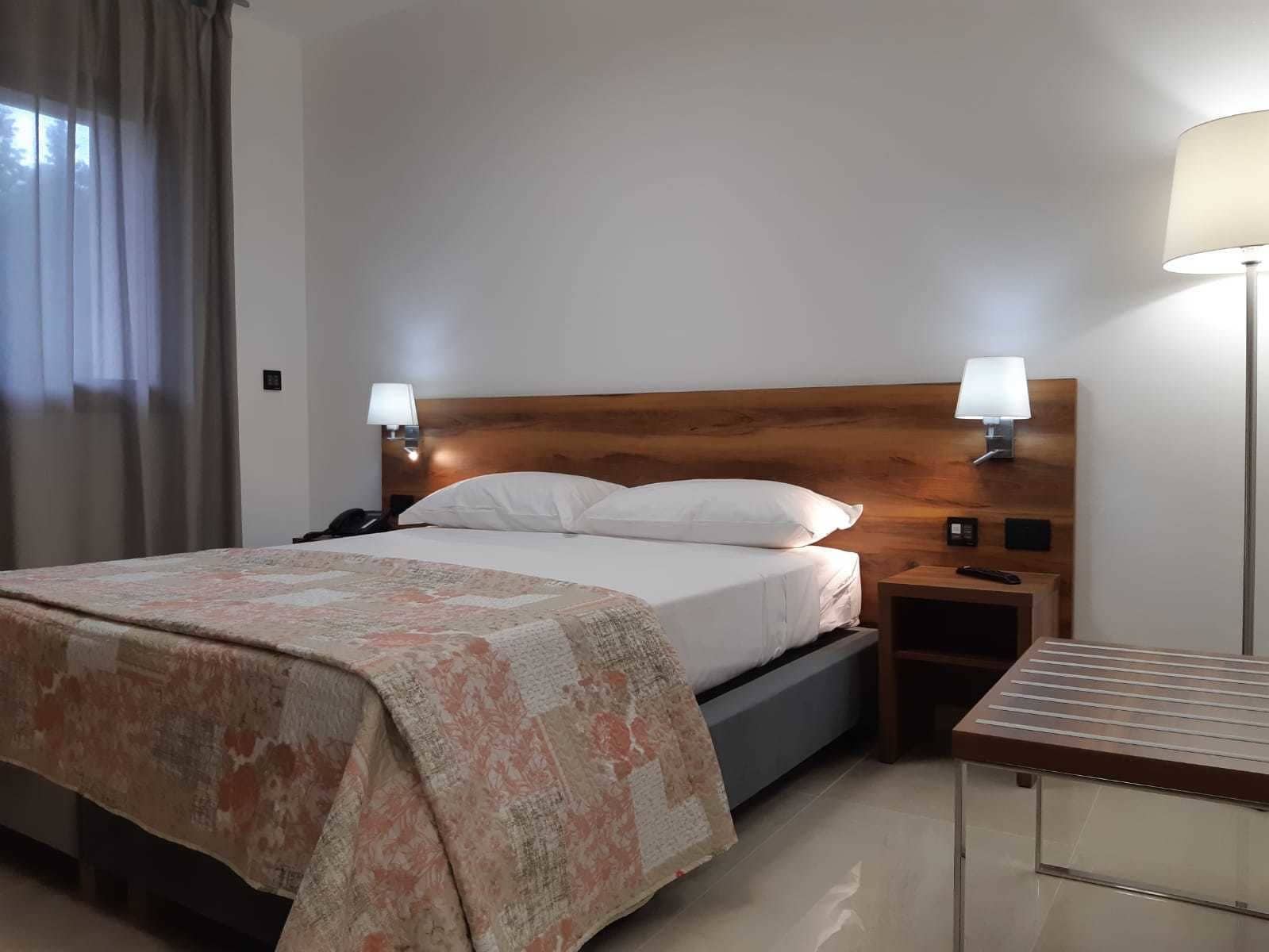 A hotel room with a wooden headboard, a bed with white linens, two bedside lamps, a nightstand, and a small table with a lamp.
