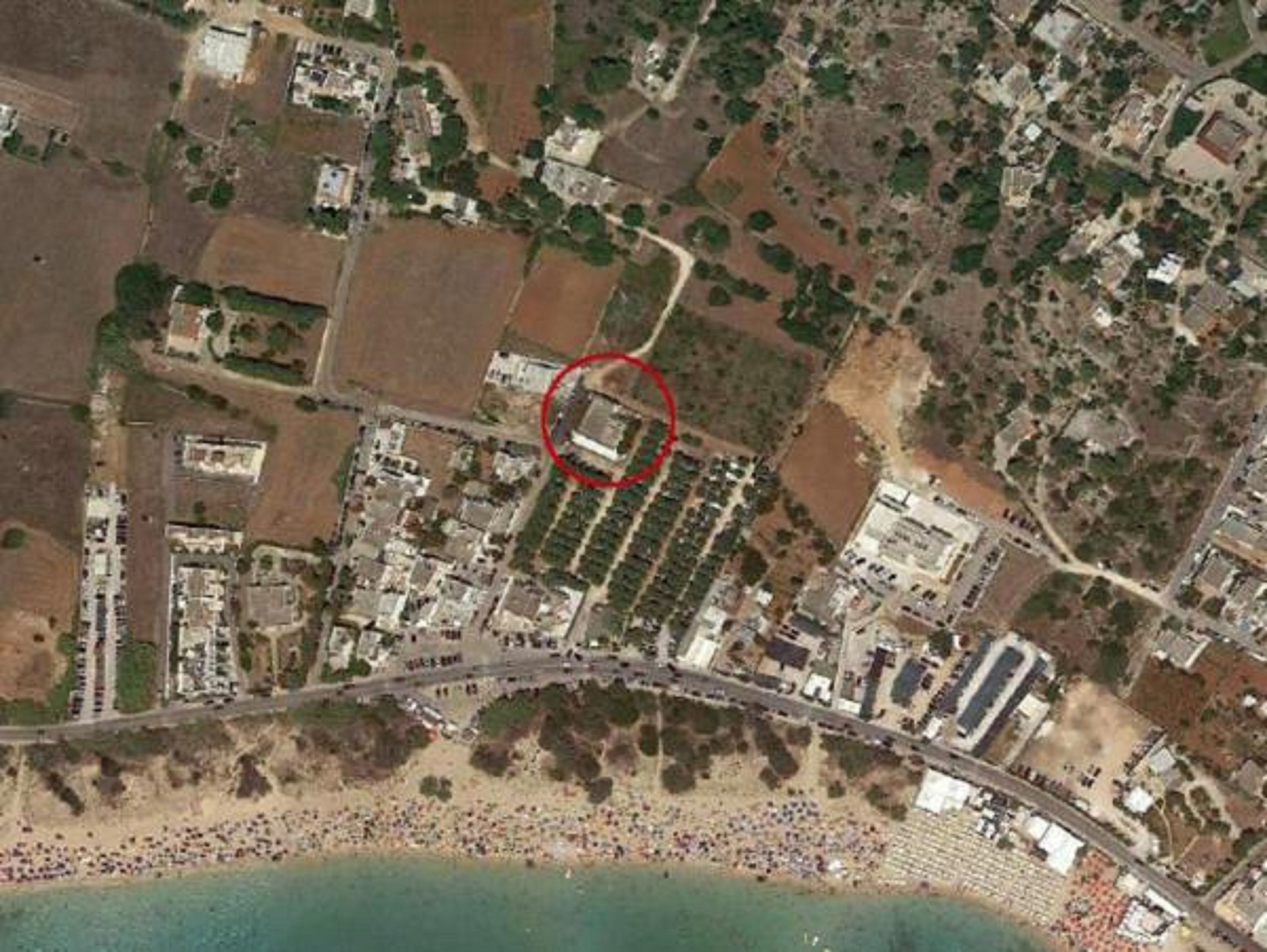 Vista aérea de un pueblo costero con playa, carretera y un complejo de edificios marcado en rojo.