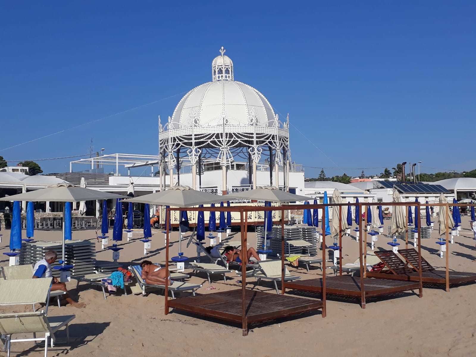 Ein Strand mit Liegestühlen und Sonnenschirmen, eine weiße Kuppelkonstruktion und ein klarer, blauer Himmel.