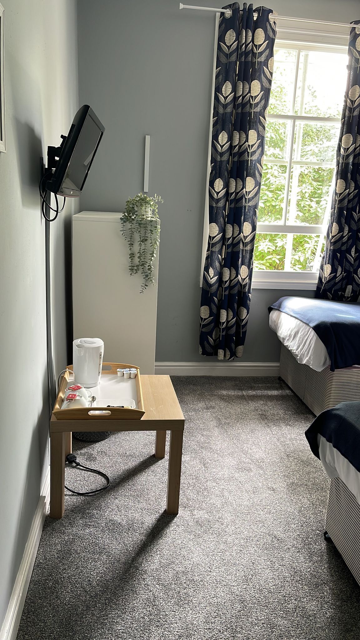 Una habitación con cama, escritorio con hervidor y tazas, televisor montado en la pared y ventana con cortinas.
