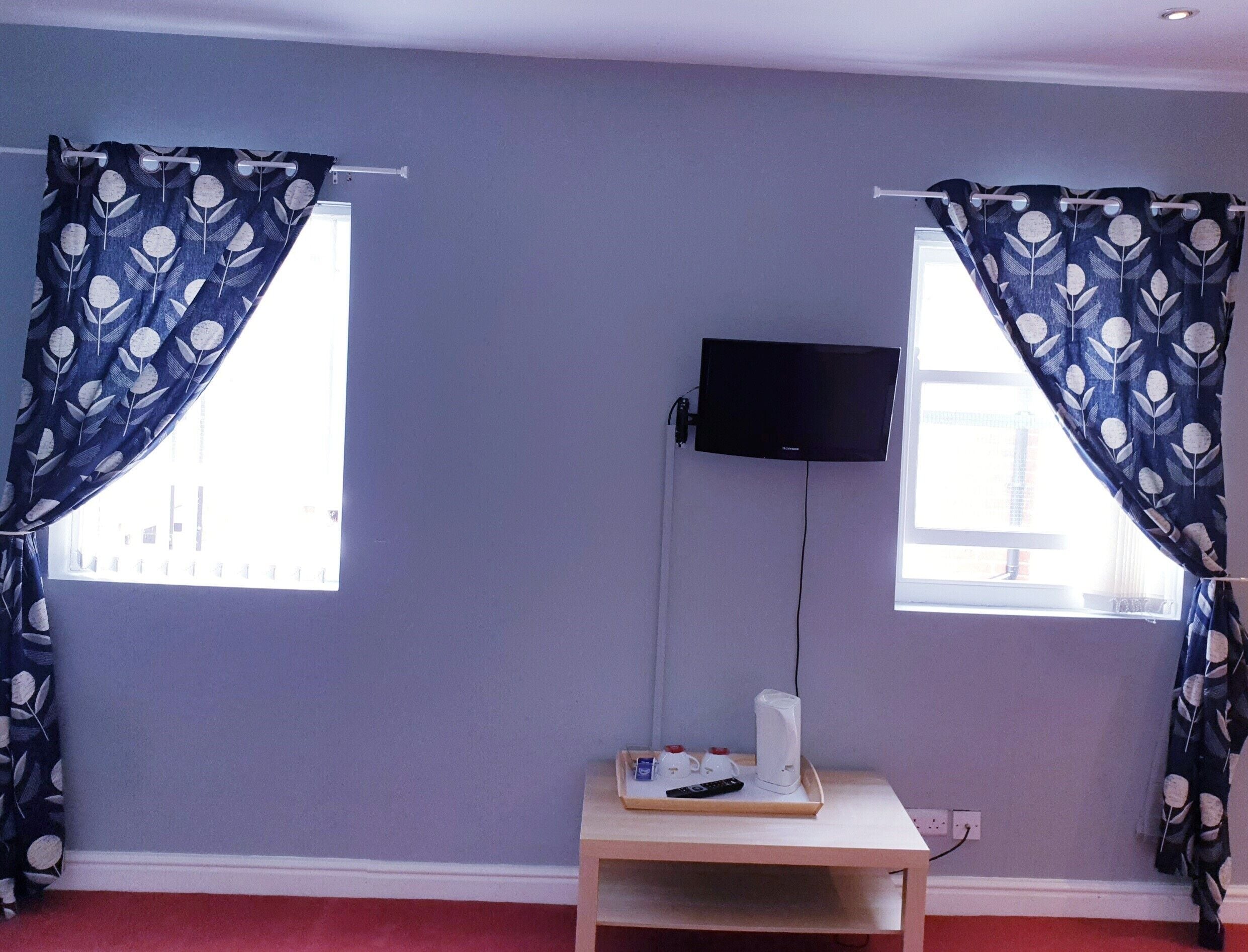 Une chambre dotée de deux fenêtres, d’un téléviseur à écran plat fixé au mur, d’une petite table sur laquelle se trouve un plateau avec une télécommande, d’une bouilloire électrique blanche et d’une prise de courant.