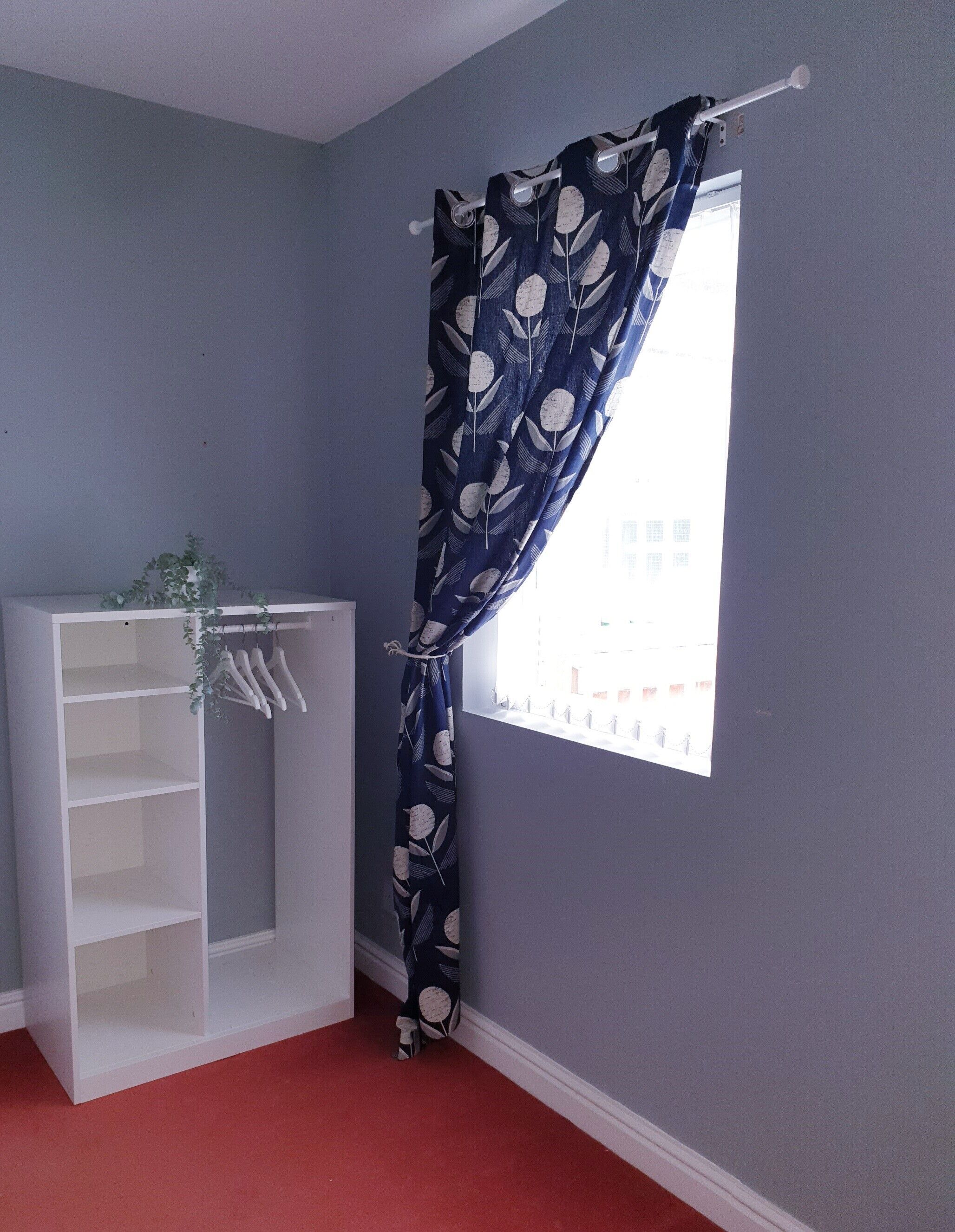 A room with a window, a white shelving unit, and a blue curtain with a leaf pattern.
