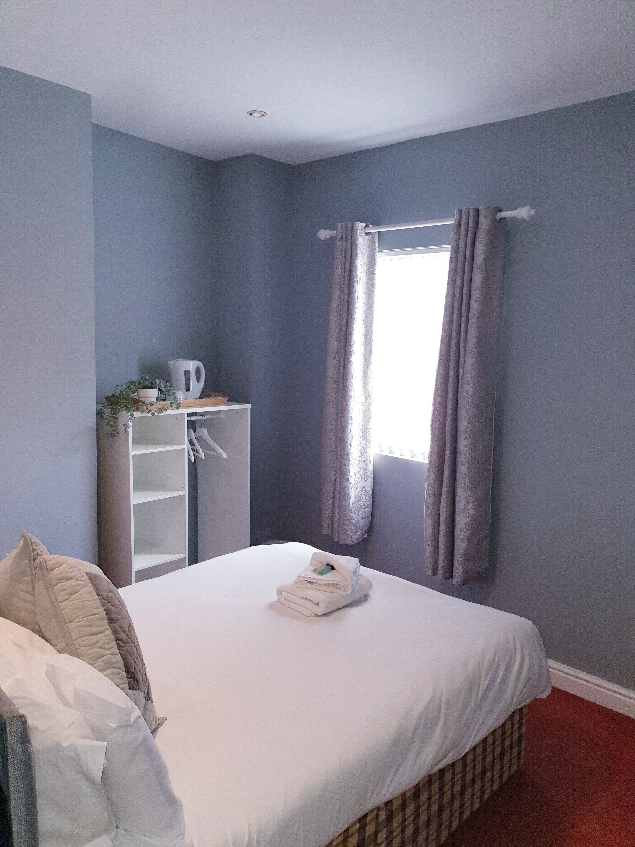 A neatly made bed with a folded towel, a white shelf with a kettle and a plant, and a window with purple curtains.