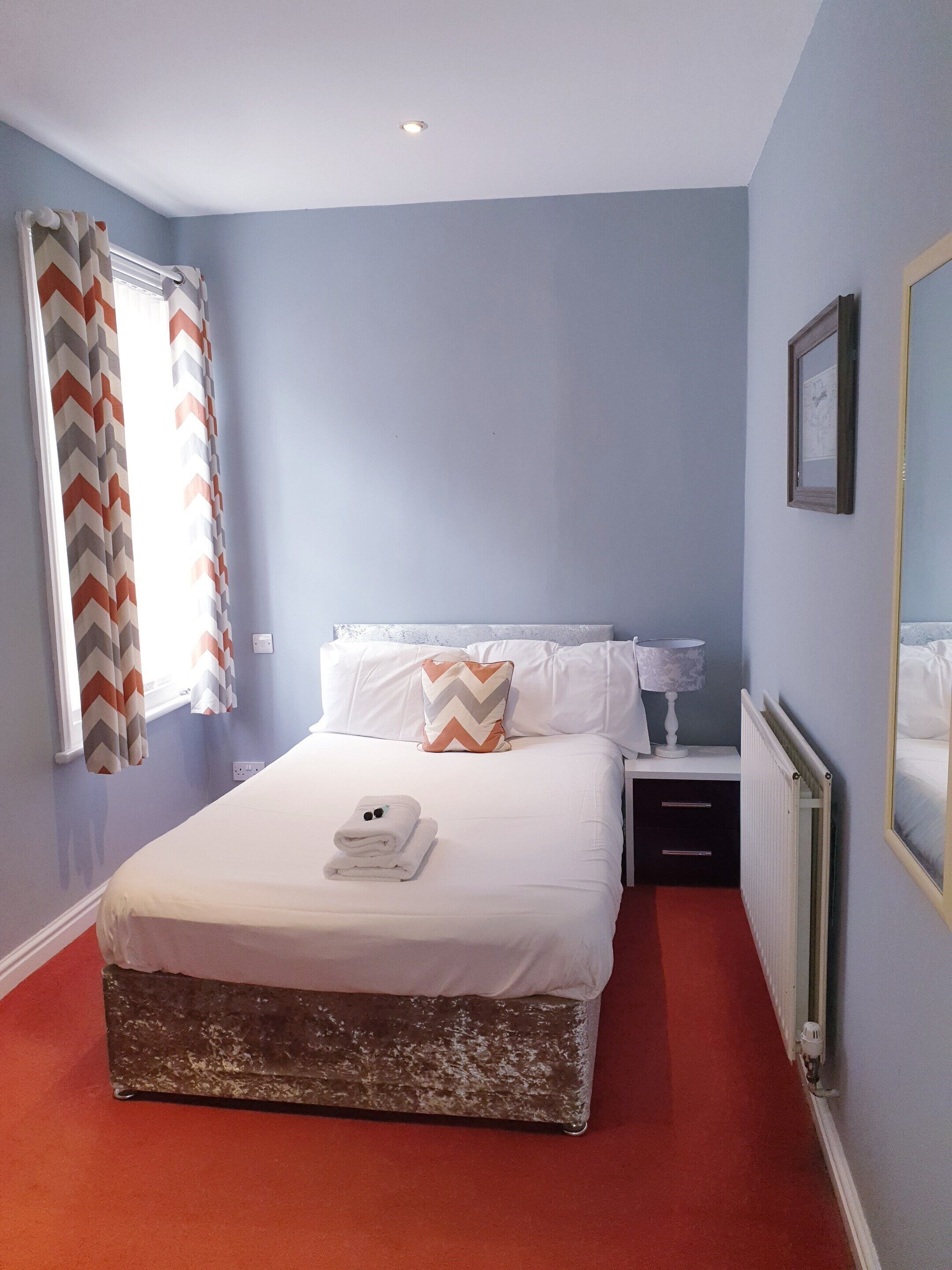 A neatly made bed with a white comforter and a folded towel, a nightstand with a lamp, a radiator, and a window with chevron-patterned curtains.