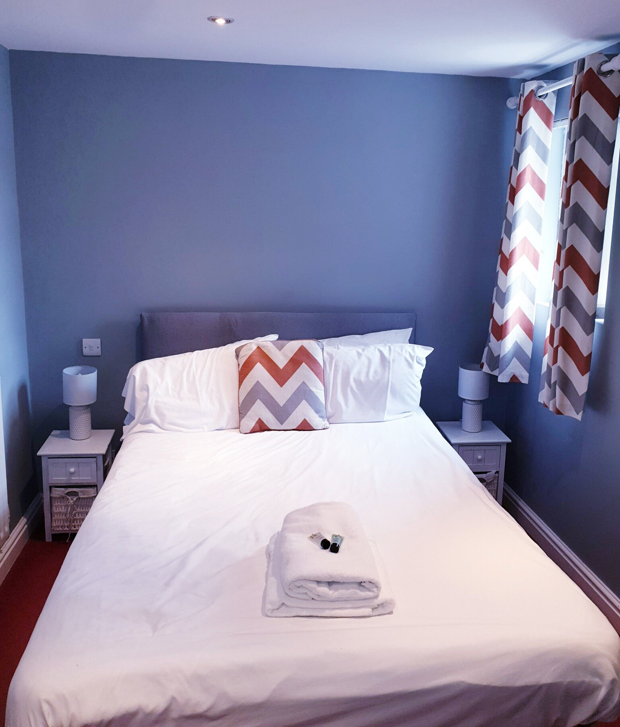 Un lit bien fait, avec du linge de lit blanc, une serviette pliée et un coussin à motifs chevrons. Il y a deux tables de chevet avec des lampes et une prise électrique au mur.