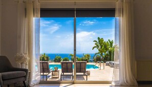 Une pièce dotée d’une grande porte-fenêtre coulissante donnant sur un espace piscine avec deux chaises longues et une table, et offrant une vue sur un ciel bleu azur et l’océan.