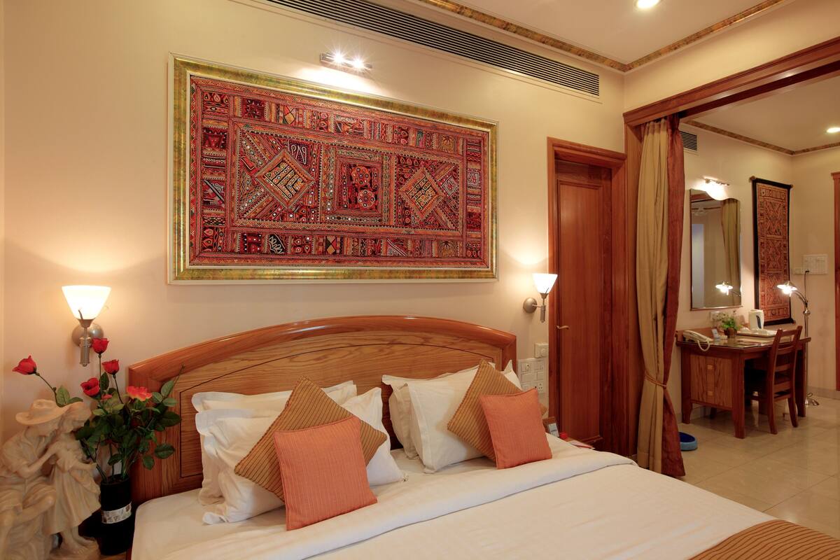 A hotel room with a large bed, a wooden headboard, a wall-mounted tapestry, bedside lamps, and a desk with a chair.