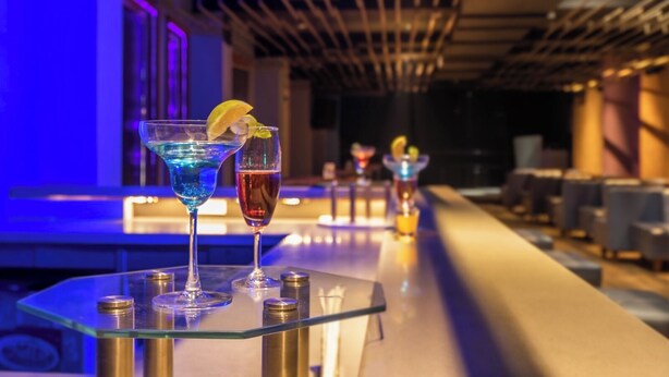A bar area with two cocktails on a glass table, a ceiling with linear lighting, and a bar counter with chairs. - Hotel Mayura (Raipur)