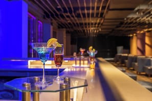 A bar area with two cocktails on a glass table, a ceiling with linear lighting, and a bar counter with chairs. - Hotel Mayura (Raipur)