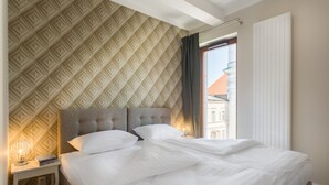 Cama bem arrumada com roupa branca, cabeceira com padrão geométrico e mesa de cabeceira com abajur e livros.