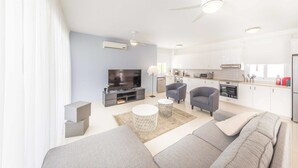 A modern living room with a sofa, a coffee table, and a TV. There is a kitchen area with cabinets and appliances. - Villa Siena (Pegeia)