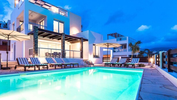 A modern poolside area with lounge chairs and umbrellas, a clear pool, and a two-story building with large windows and a balcony. - Villa Siena (Pegeia)