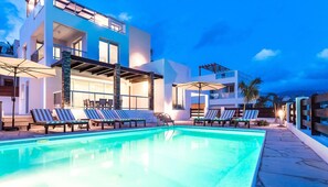 A modern poolside area with lounge chairs and umbrellas, a clear pool, and a two-story building with large windows and a balcony. - Villa Siena (Pegeia)