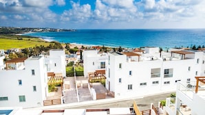A coastal residential area with modern white buildings, a clear view of the ocean, and a paved walkway leading to a wooden pergola. - Villa Siena (Pegeia)