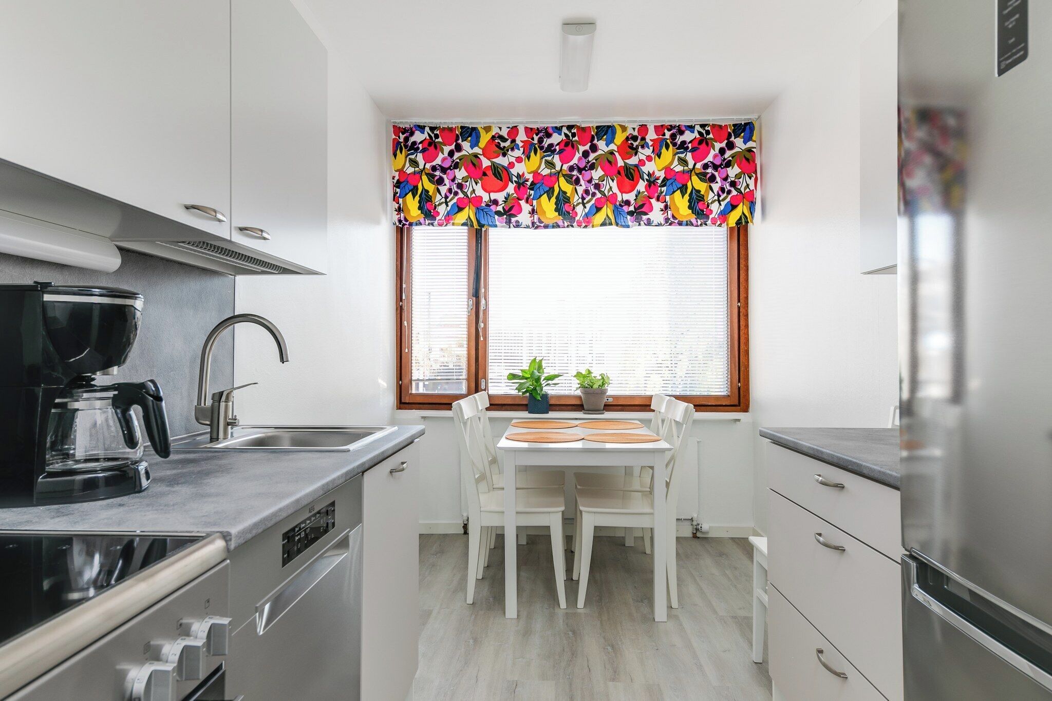 Uma cozinha moderna com armários brancos, geladeira de aço inoxidável, cafeteira e uma janela com cortinas coloridas.