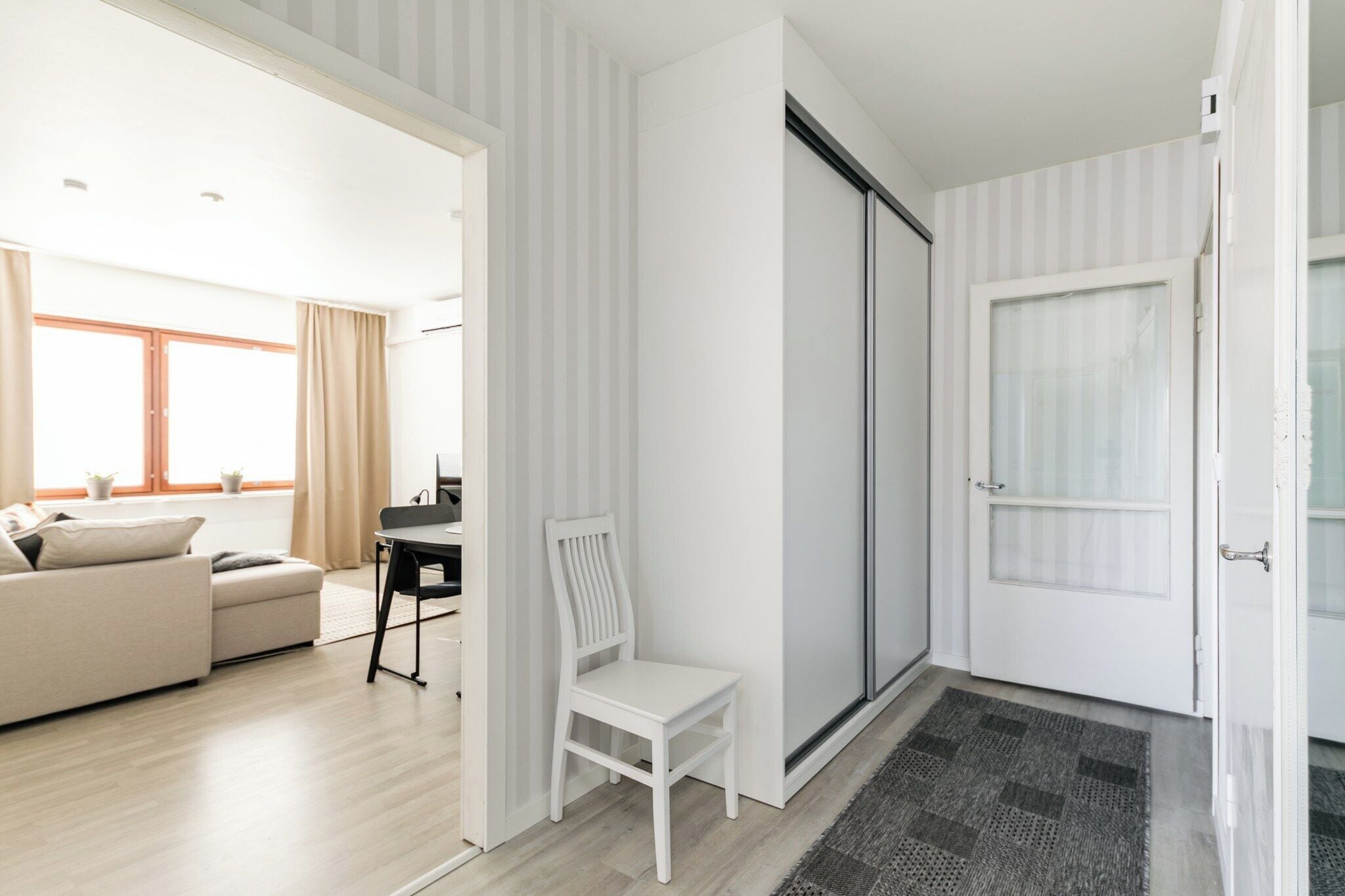 A modern living room with a sofa, a dining table, and a chair. There is a sliding door and a window with curtains.