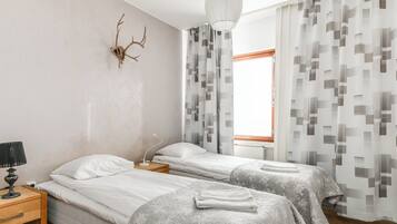 A hotel room with two beds, a nightstand with a lamp, a wall-mounted deer head decoration, and a window with patterned curtains.