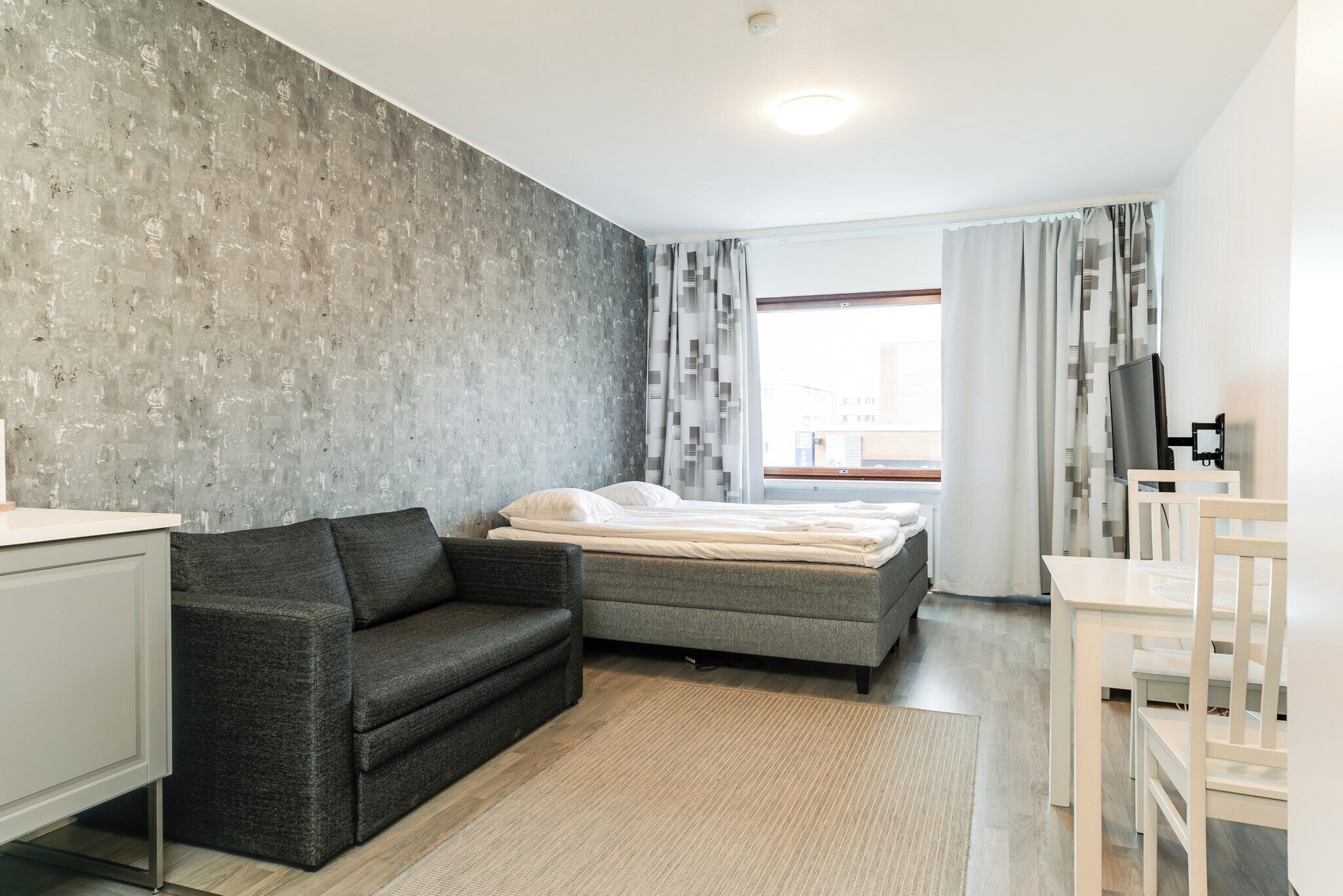 A modern bedroom with a grey sofa, a bed with white linens, a small table, and a TV.