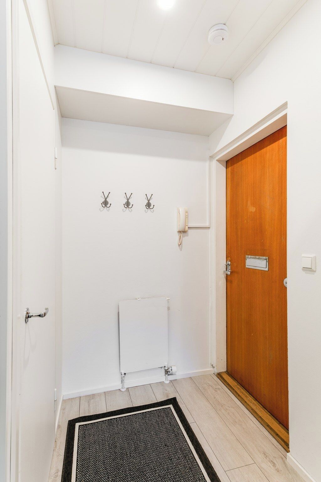 Un pasillo con puerta de madera, un radiador y tres ganchos en la pared.