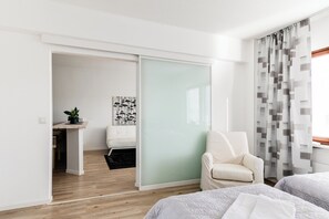 A modern hotel room with a sliding door leading to a bathroom, a white armchair, two beds, and a view of the outside through a window with patterned curtains. - Santa's Holiday Homes (Rovaniemi)
