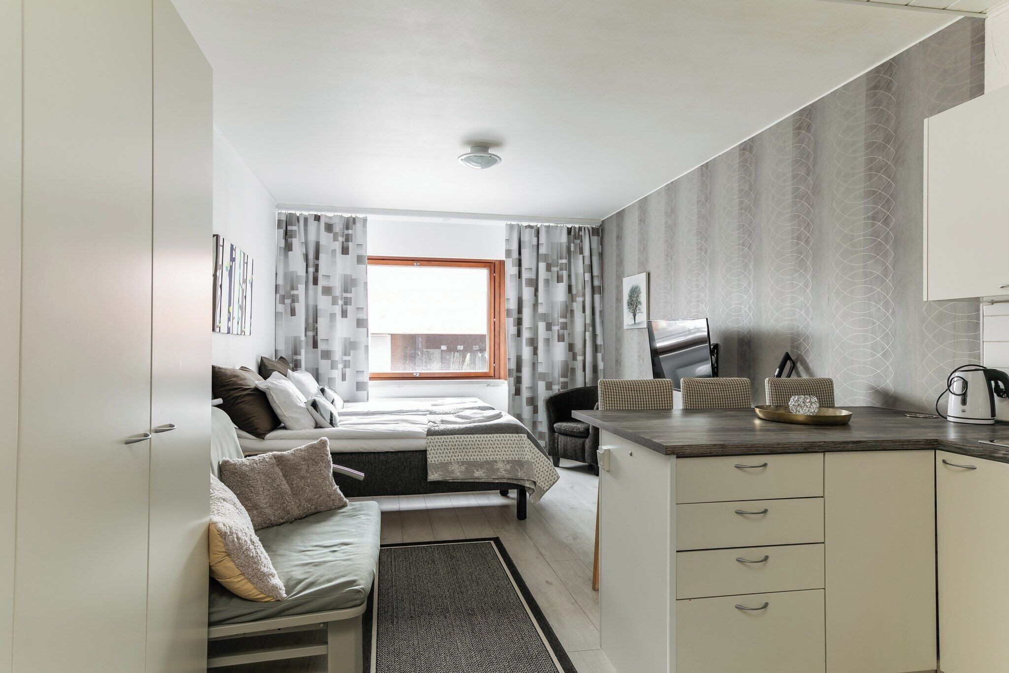 Une chambre compacte comprenant un lit, une table de chevet et un petit coin salon. On peut apercevoir une kitchenette équipée d’une cuisinière et de placards.